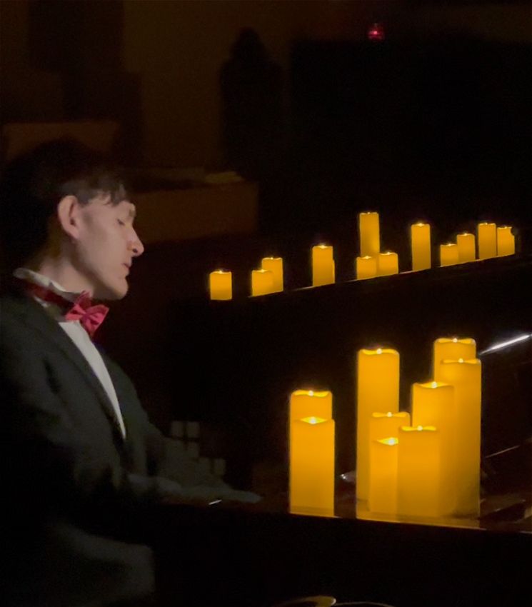 A photo that appears to be taken mid-performance where Matthew seems focussed and impassioned as he plays the piano. They room around them is dark, but the scene is lit with large spherical candles of varying lengths, of which we 7 rest atop the piano and the rest are scattered around the room. Matthew is wearing a black suit with a white shirt and a red bowtie.