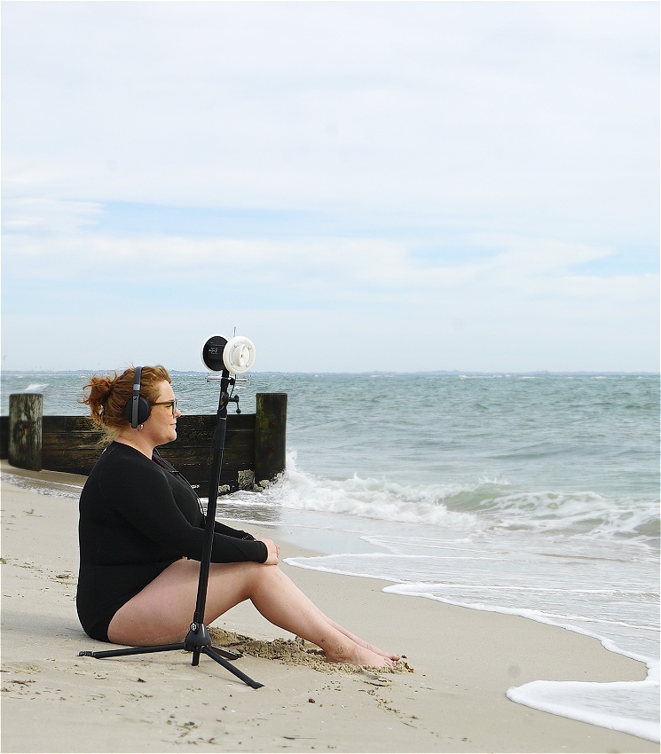 A young woman wearing a black wetsuit sits on a beach looking into the ocean. She’s wearing black headphones over her messy bun. Next to her is a tripod with a microphone on top of it, and the microphone looks like it has two human ears on either side of it, but they’re made out of a white plastic. The waves are almost lapping at here feet and the ocean looks calm and turquoise.
