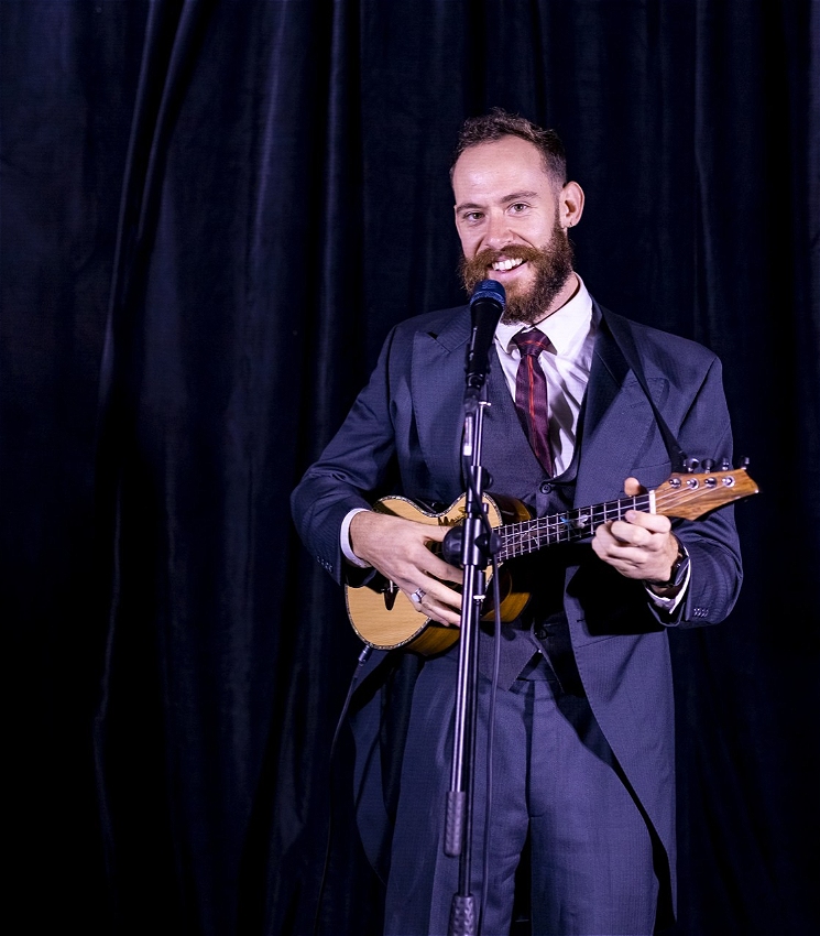 30 y.o bearded man in snappy navy suit and burgandy tie, holding a ukulele on stage in front of microphone with a cheeky smile