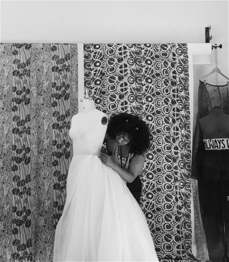 A fashion designer works delicately pinning fabric to a mannequin bust to create an elegant gown. There are two panels of fabric behind them that have intricate patterns on them.