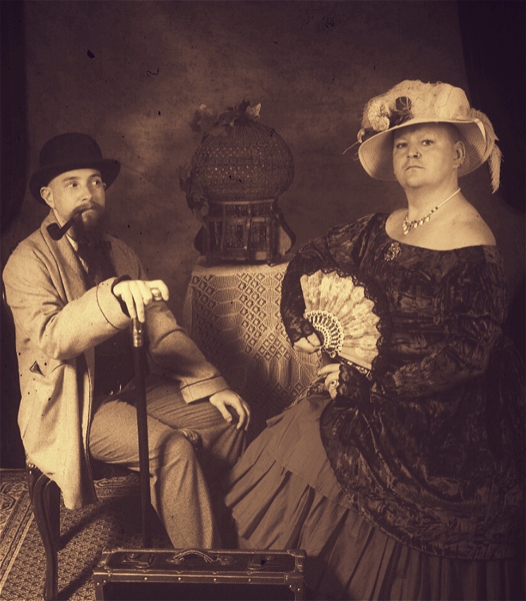 A sepia photograph with a couple in vintage style clothes sitting in a living room setting. One, a bearded man in a loose suit looks with suspicion at the other, a nonbinary person looking at the camera with no hair or eyebrows in a full dress and hat ensemble holding an open fan.