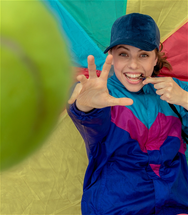 A woman wearing a blue cap and a blue and purple puffy jacket. She is smiling and holding a whistle to her mouth with her right hand. Her left hand reaches forward to catch the tennis ball that is also in the frame.