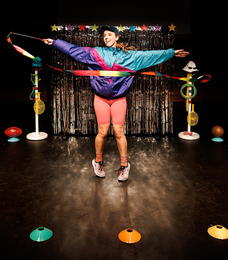 A woman wearing a blue cap, a blue and purple puffy jacket and pink shorts holding a dance ribbon in motion. She is standing on a dark stage in front of a silver curtain that is shimmering. She is smiling and has her arms stretched out.