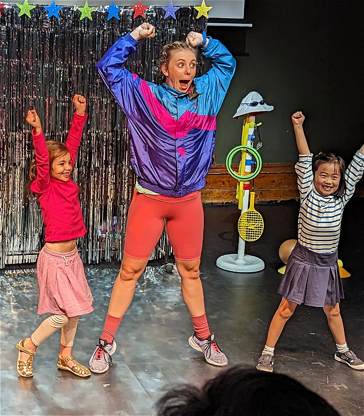 A woman wearing pink and purple is standing with her arms in the air as if celebrating. She has two young girls next to her who are also doing the same celebratory action. They are standing on stage in front of a silver curtain.