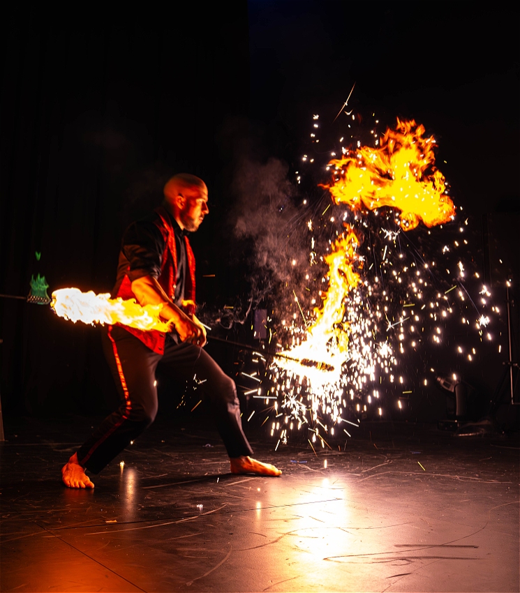 In a dark room with a black floors, a performer is spinning a lit flower stick that emanates strong golden flame on both ends. The performer is barefoot and dressed in a black shirt and trousers with a red waist coat. There's also a burst of sparks in front of them, which they stare at intently, as its reflection creates a large flare of flight on the floor.