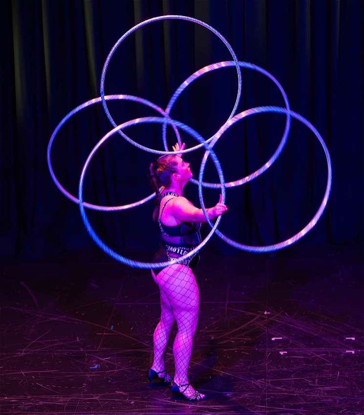 a performer is standing on a dark stage. balancing a blue hula hoop on their head while juggling 4 more in their hands. there is pink hue on the performer, they are wearing fishnets, and a tight black top .