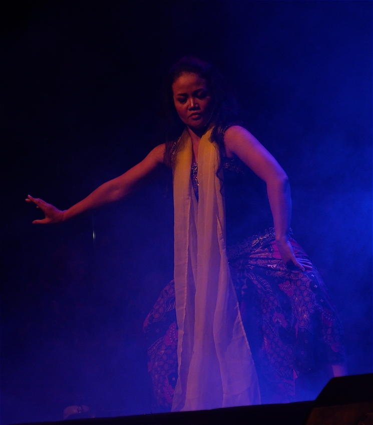An Indonesian woman is standing in the middle of the picture in a dance pose looking down. She is wearing a long white scarf with a dark singlet top and patterned pants. The image is theatrical with haze with blue and purple lighting.