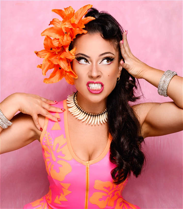 A Hawaiian woman with long dark hair and huge orange flowers in the right side of her hair standing in front of a pink furry backdrop. Wearing a neon pink Pasifika inspired dress with neon orange hibiscuses and traditional Hawaiian patterns on it. Looks up to her left