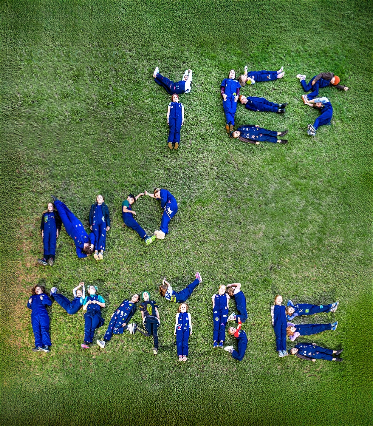 A birds eye view of 27 children wearing navy overalls lying on green grass, using their bodies to spell out the words: YES, NO, MAYBE.
