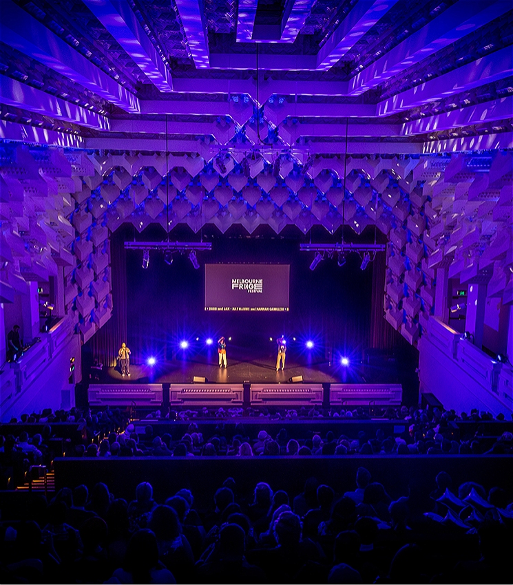 A crowded theatre packed with people watching two performers on the stage. There is a projector above the performers with text that reads: Melbourne Fringe Festival. The lighting is dramatic and blue.