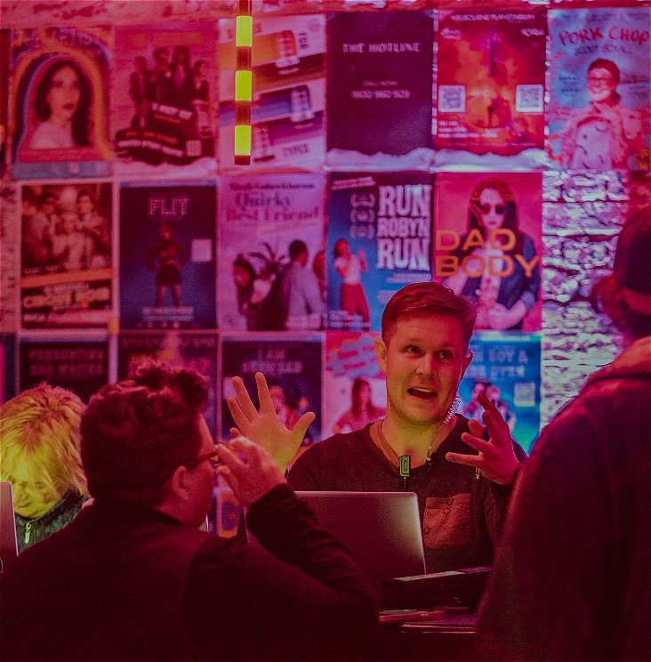 Someone from box office stands in front of a colourfully lit wall of event posters. They are gesturing with their hands like something is large and exciting.