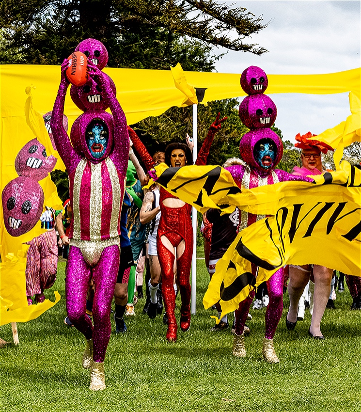 Dozens of people dressed in sparkly and glittery costumes run through a sports banner on a grassy field.