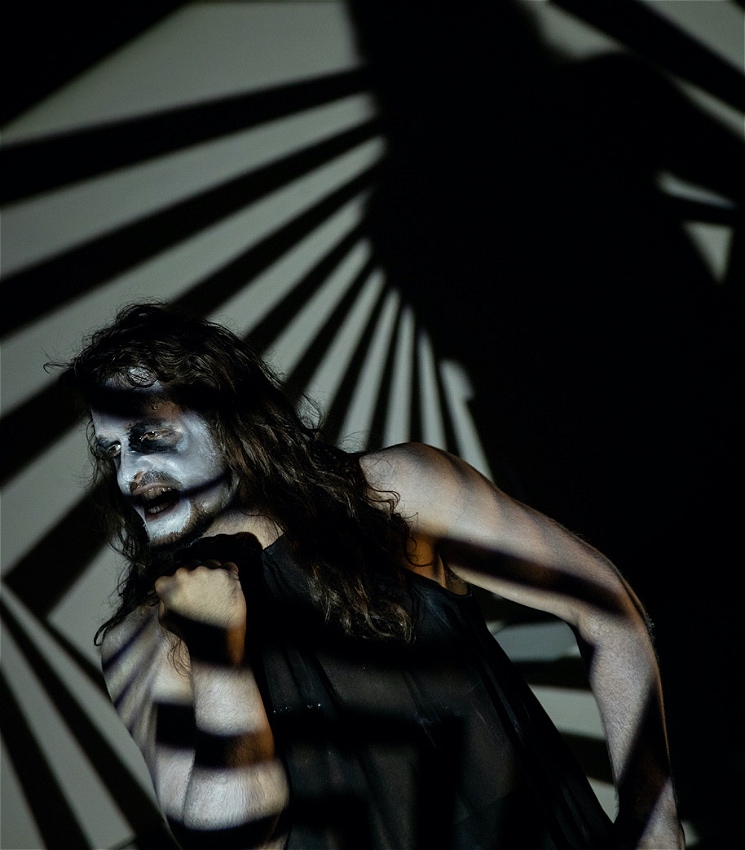 A ghoulish figure in black with a white made up face looks mysteriously away from the cameras. The lighting suggests barred windows in different orientation.
