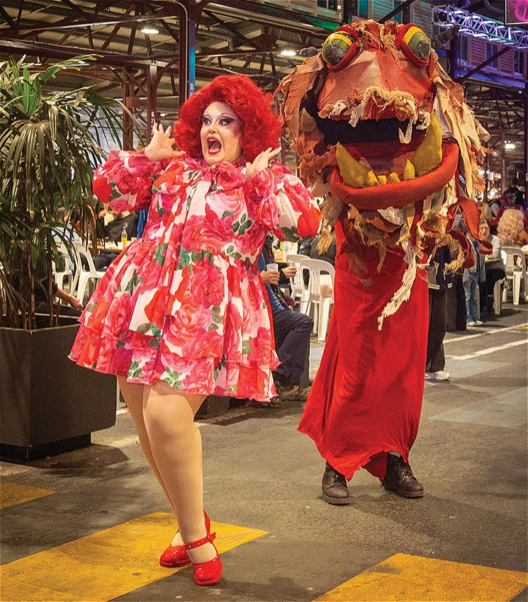 A drag performer in a vibrant red wig and a bold floral dress strikes an animated pose with wide eyes and hands raised, standing next to a large, playful red monster puppet with exaggerated yellow teeth and bulging eyes. They are in a lively indoor venue filled with people, plants, and rows of white chairs under industrial-style lighting and signage.