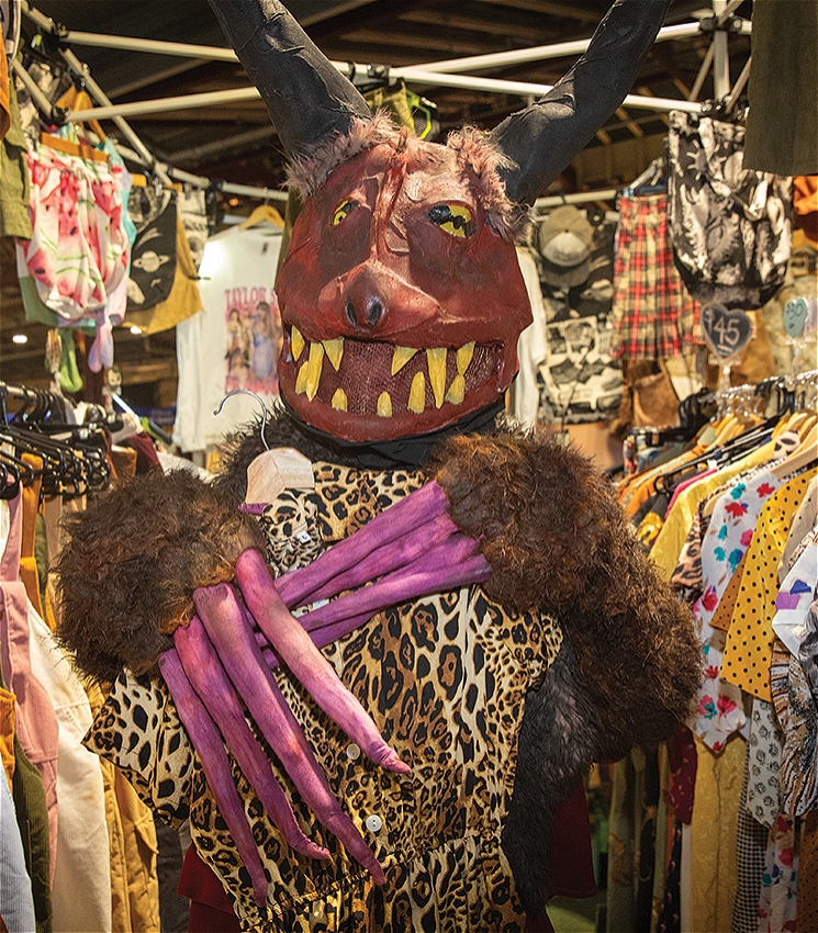 A whimsical monster character with large black horns, yellow teeth, and purple claws poses in a vintage clothing stall, holding up a leopard-print dress. The creature is surrounded by colorful racks of secondhand clothes, including dresses, overalls, and patterned shirts, with hand-written price tags visible. The playful scene takes place indoors under market-style lighting.