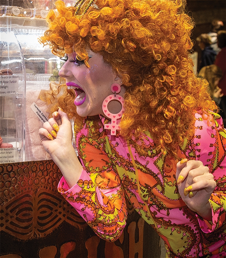 A performer with bright orange curly hair and bold makeup excitedly peers into a confectionery display. They wear a vibrant pink and orange patterned outfit, large pink earrings shaped like the female symbol, and matching yellow nail polish. Their expression is joyful and animated, surrounded by a festive indoor atmosphere.