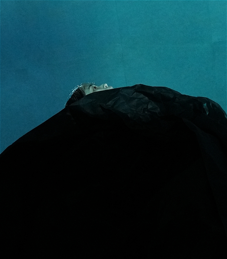 A dramatic image of a performer wrapped in a large black piece of paper, they look thoughtfully skywards. Behind the performer is a deep blue backdrop.