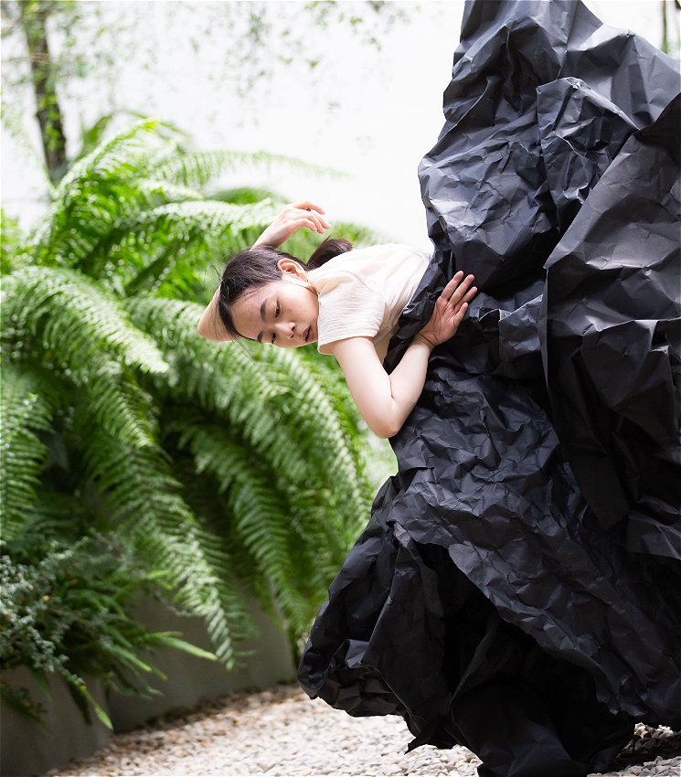 A dancer leans gracefully to the side with eyes closed and arms curved overhead. They wear a beige top and are partially wrapped in a large, sculptural sheet of crumpled black paper. Behind them, lush green ferns and trees frame the scene, with pale pebbles underfoot.