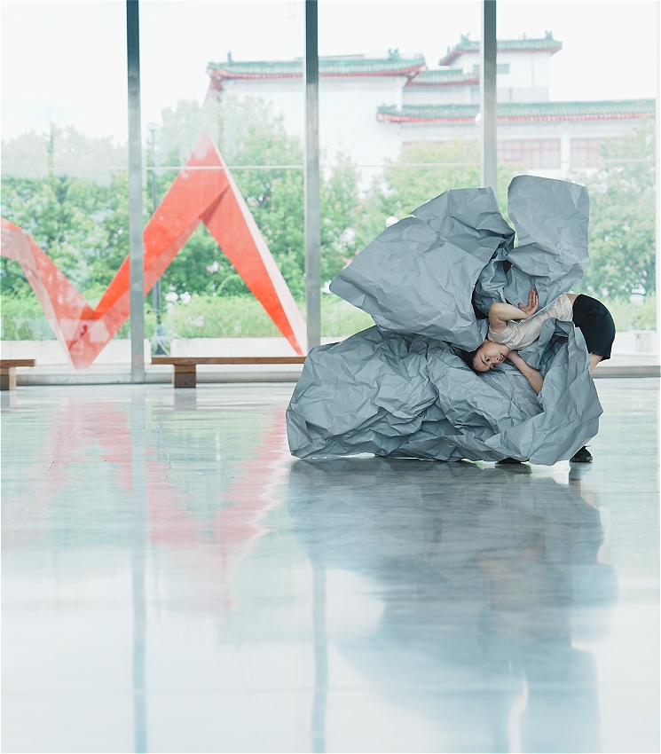 A dancer bends low to the ground, partially enveloped in a large, sculptural sheet of crumpled light blue paper. They wear a beige top and black shorts. The scene is set in a spacious, reflective hall with floor-to-ceiling windows. Outside, greenery and traditional-style buildings are visible, with a bold red geometric sculpture seen through the glass.