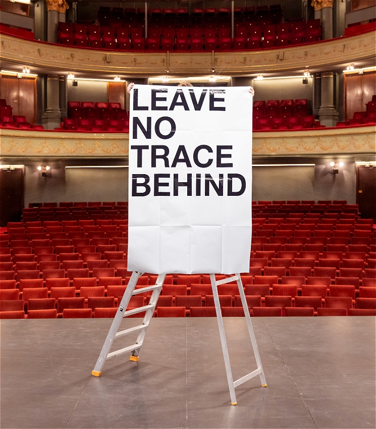 A white ladder on stage with a large white piece of paper that reads: Leave No Trace Behind. There is a person behind the paper who is barely visible. The photo is taken from the perspective of looking out to the auditorium, and it is a large theatre with a dress circle and balcony in view, with lush red velvet seats.