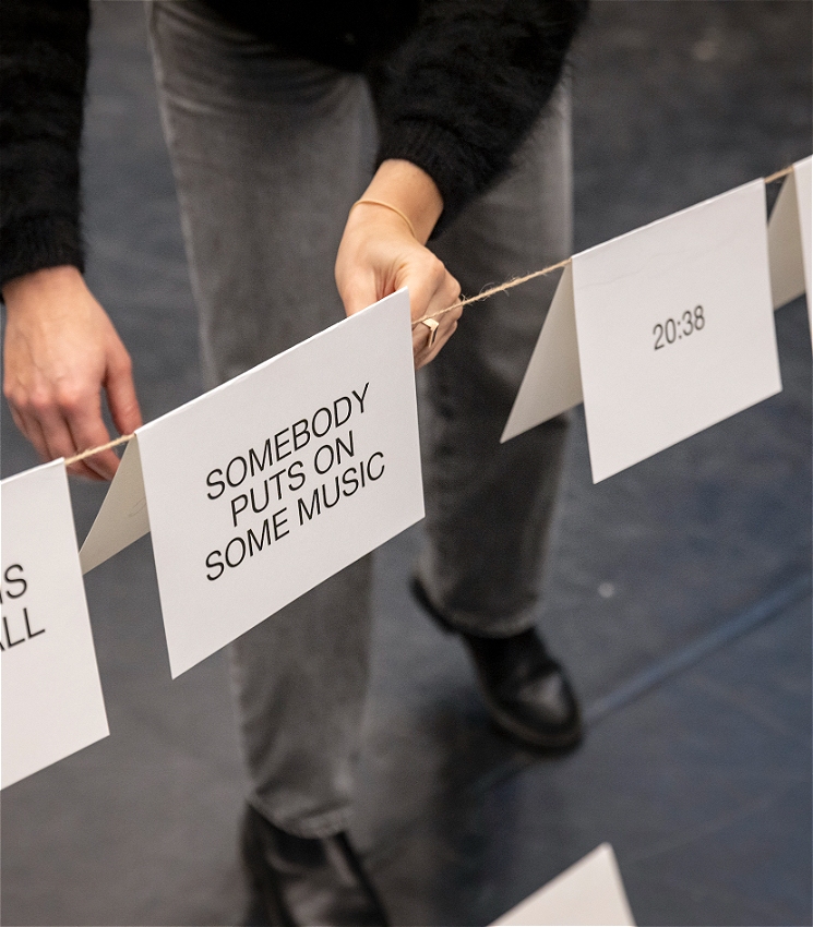 There is string suspended across a room with folded cards hanging over it. A person stands behind it, out of focus and places a new card on the string. The new card reads: Somebody puts on some music.