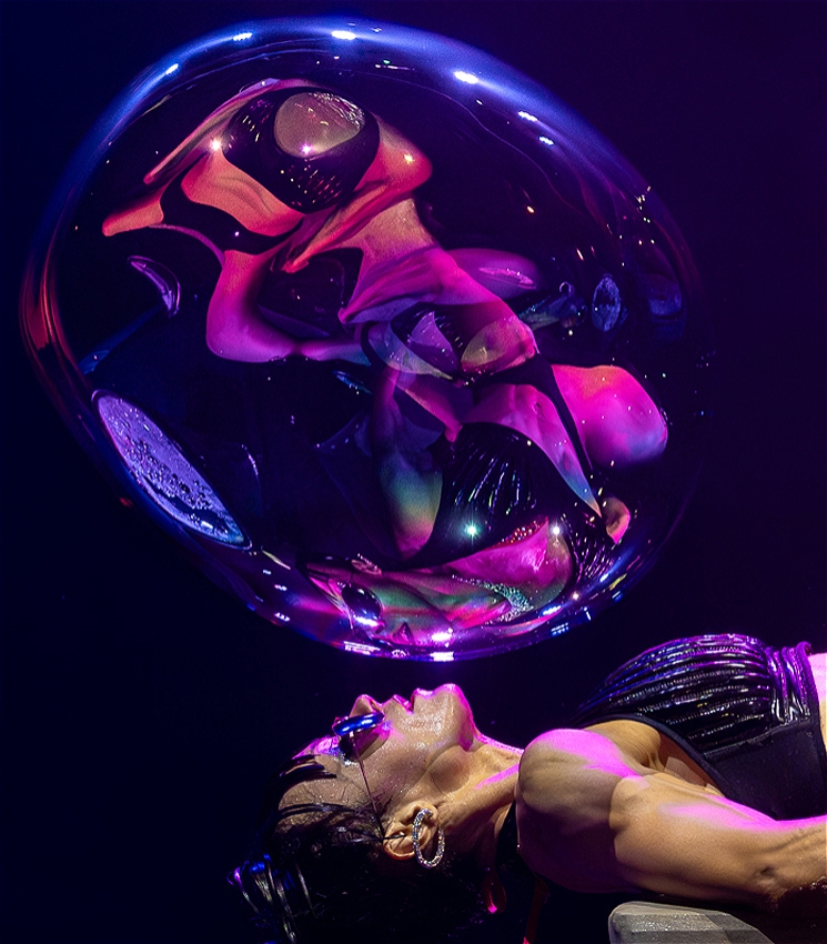 A woman in a leather bikini watches a giant bubble filled with her reflections float above her face