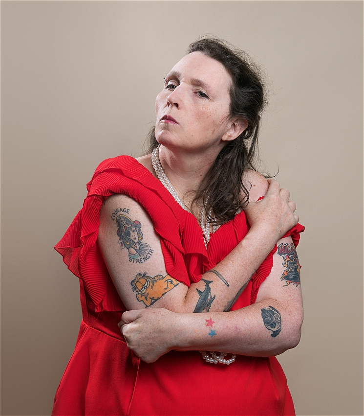 Louise, a white woman with dark hair and an eyebrow piercing, pouts her lips and looks off into the distance. She wears a frilly red gown and long strands of pearls. Her arms are crossed over her chest and they are covered in tattoos: a shark, a pirate, Garfield, a pug, and the AC/DC logo.