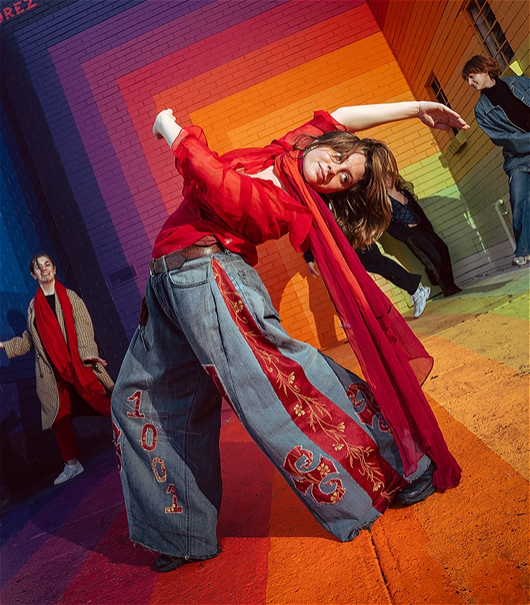A dancer wearing denim jeans and a red top in the middle of the image bends backwards with their arms outstretched. There are four other dancers in the background. All of the dancers are in front of a brightly coloured painted rainbow mural by Drez.
