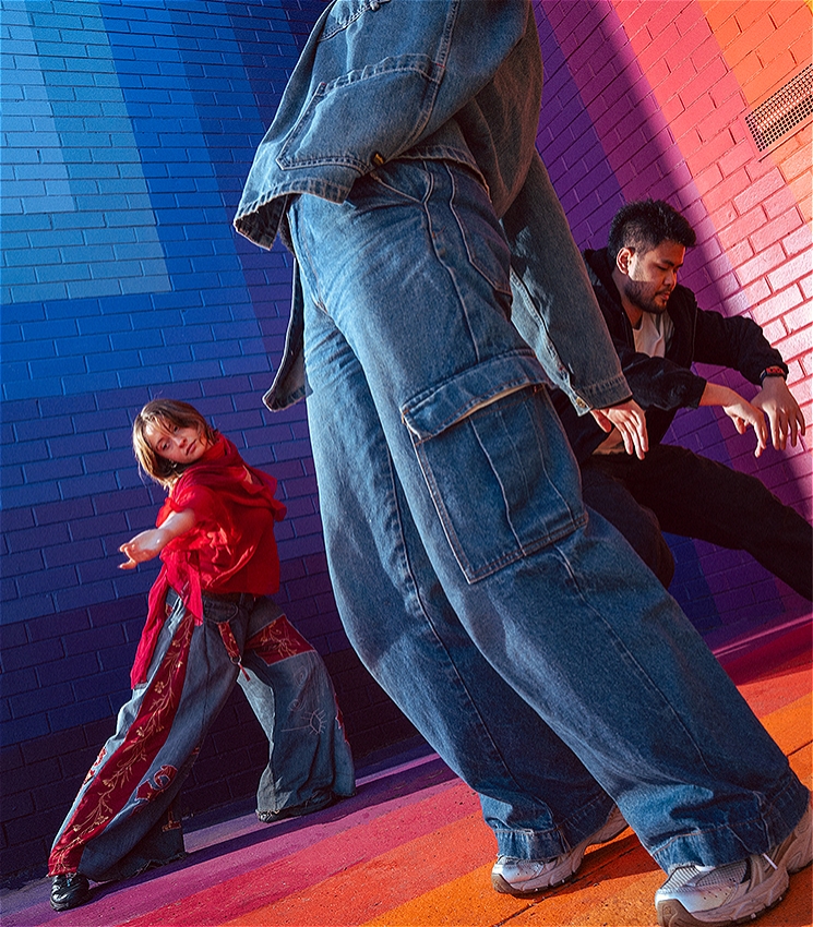 One dancer wears all denim and strikes a pose in the middle of the image. There are two other dancers moving behind them. All of the dancers are in front of a brightly coloured painted rainbow mural by Drez.