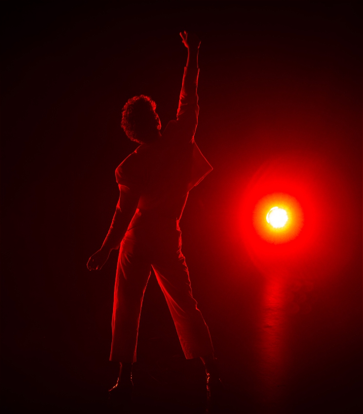 A performer stands on a dark stage with one arm stretched above their head. They are backlit by a bright red light.