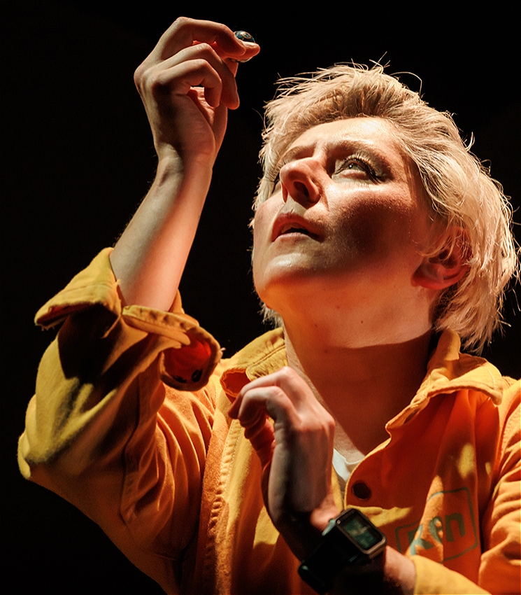 A person in a mustard yellow boilersuit holds a small ball in their hand and peers up at it with a quizzical expression.