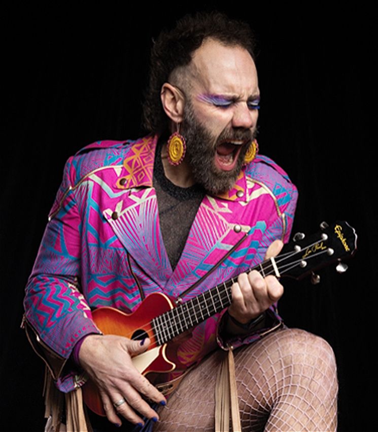 a close up photo of a person with a colourful jacket, fishnets and big dangly earings. they are holding a ukelele and poking their tongue out in a kiss style rock pose