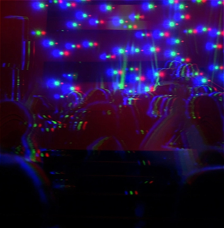 A crowd dance in a large ballroom. There is a mirror ball on the ceiling and the room is lit with purple-hued party lights.