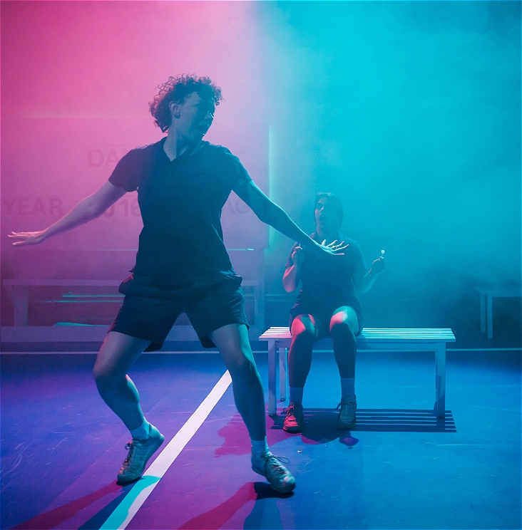 Two performers on stage in pink and blue light with haze around, they look surprised and energetic