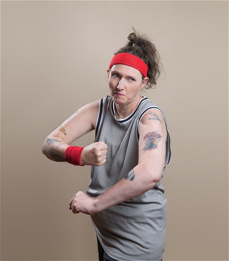 Lou wears a grey basketball jersey and red sweatbands poses in a mock-tough stance, fists raised. She has multiple tattoos on both arms, including bold text and cartoon designs. Her hair is tied back with a red headband.