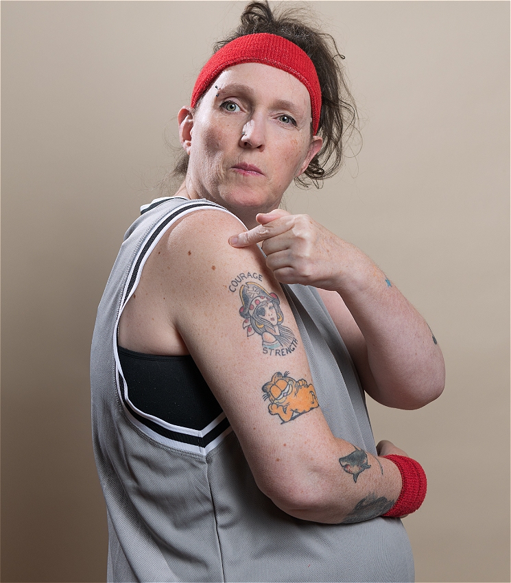 Lou wears a grey basketball jersey, black sports bra, and red sweatband on their head and wrist points to a tattoo on their upper arm. The tattoo depicts a cartoon-style mouse wearing a helmet, with the words “Courage” above and “Strength” below. Another tattoo of Garfield the cat is visible on their forearm, along with smaller tattoos. Lou looks directly at the camera with a cheeky expression.