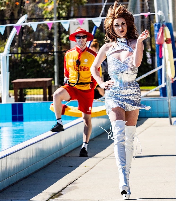 A performer with long brunette hair walks next to a swimming pool, wearing a short silver dress and over-the-knee silver boots. There is a lifeguard out of focus, smiling and standing behind them, with one foot resting on the edge of the swimming pool.