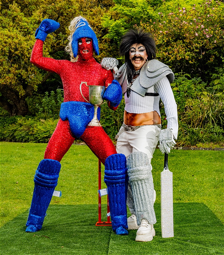 A picture of 2 performers standing on green grass with green trees behind them. The performer on the left is dressed in a fully bright red sequinned outfit and with royal blue sequinned boxing gloves, helmet and briefs, their right hand is raised with a victory fist. The performer on the right is wearing a silver cricket outfit with cricket leg pads on, with a cricket bat in their left hand. They are both smiling.