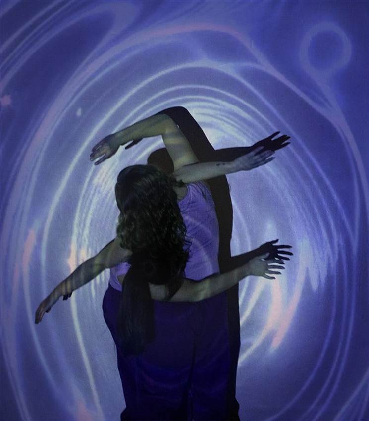 Two women are intertwined with their arms around each other. There are blue water link ripples projected over their bodies and the background.