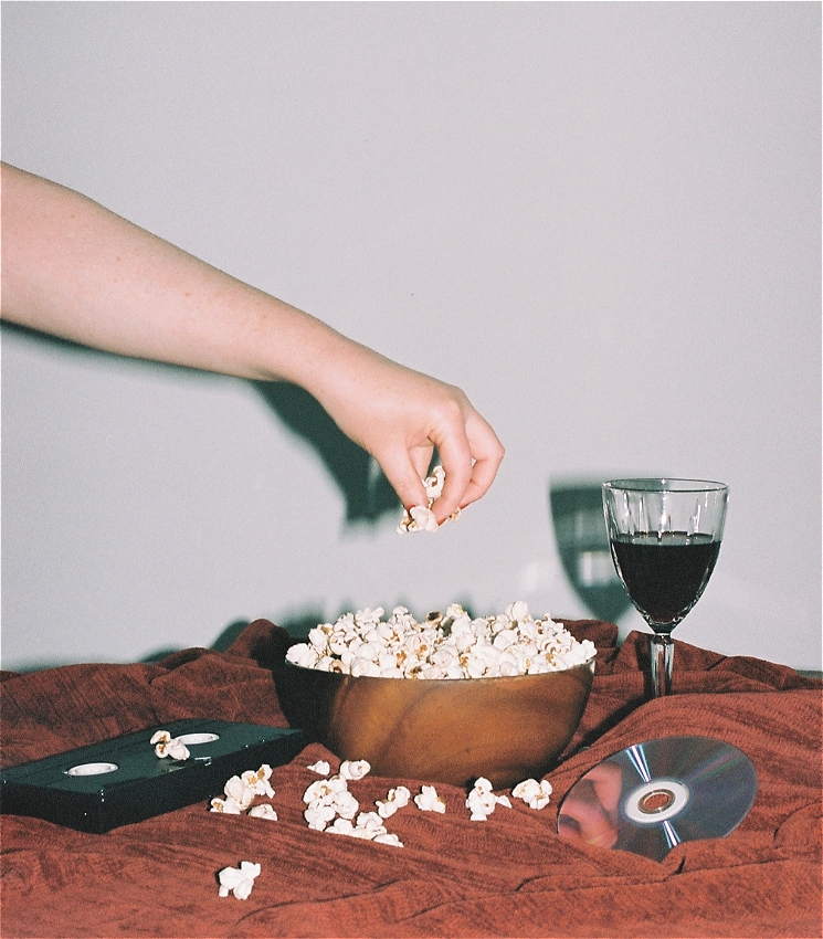 A similar flash photo taken with a film camera.
In the middle of the shot is a bowl of popcorn and a hand that has just grabbed some the popcorn. The bowl is on a table with a red tablecloth.
On the left of the bowl is a black VHS, and on the right of the bowl is a retro crystal wine glass with red wine and a blue reflective DVD.