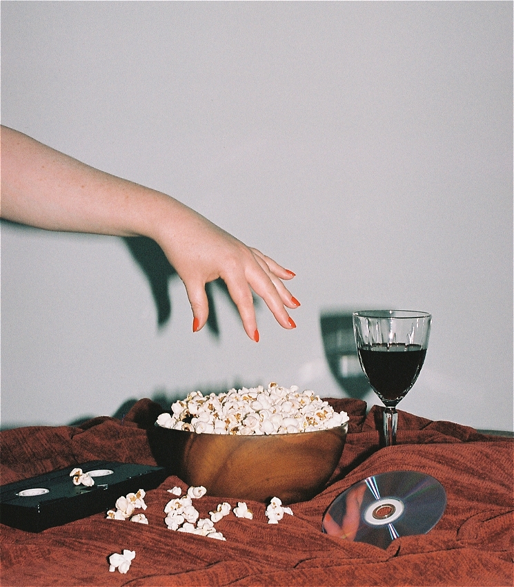 A flash photo taken with a film camera.
In the middle of the shot is a bowl of popcorn and a hand letting go of some popcorn it may have previous grabbed. The bowl is on a table with a red tablecloth.
On the left of the bowl is a black VHS, and on the right of the bowl is a retro crystal wine glass with red wine and a blue reflective DVD.