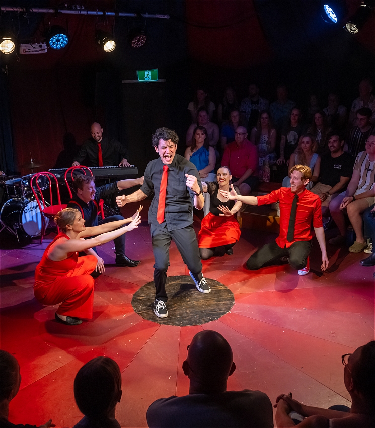 A male performer singing his guts out with surrounding cast forming levels around him. There is a smiling audience in a tent.