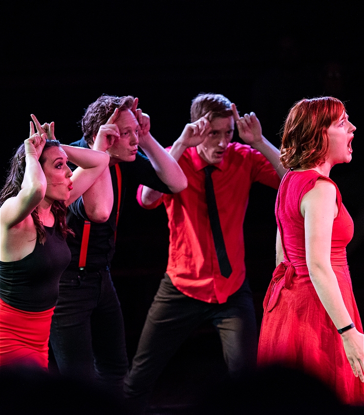 One female singer facing right and three cast members committedly being cows as backup. All are dressed smartly in red and black.