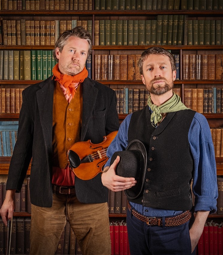 Two white man stand in front of a bookshelf staring sternly at the camera. Sammy on the left is wearing a blue shirt and a black coat with a green scarf and is holding a black hat. To his left the person is wearing a red shirt with a orange waistcoat and neck scarf with a black jacket - his also holding a violin and it's bow.