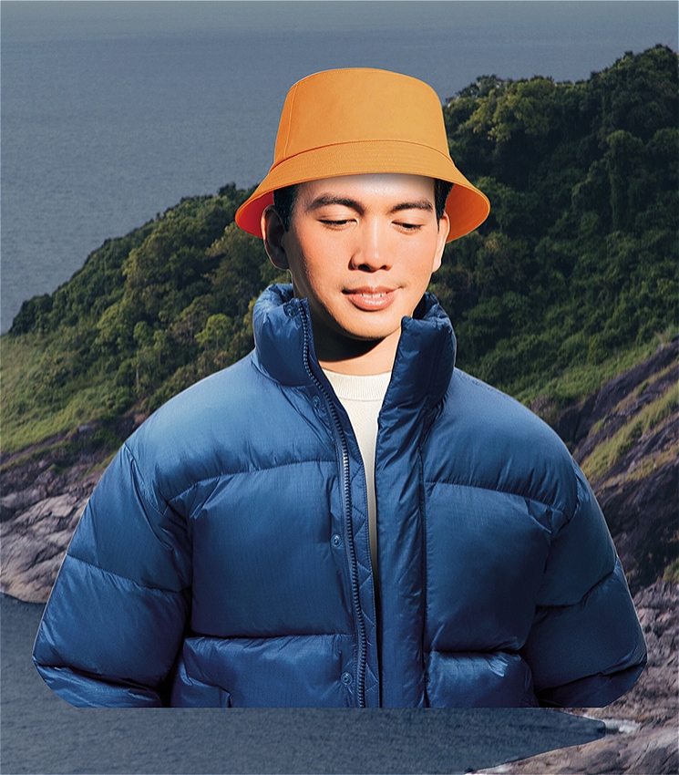 A naive-looking character wears an orange bucket hat and a blue puffer jacket. He has a slight smile. In the background is a tropical coastline.