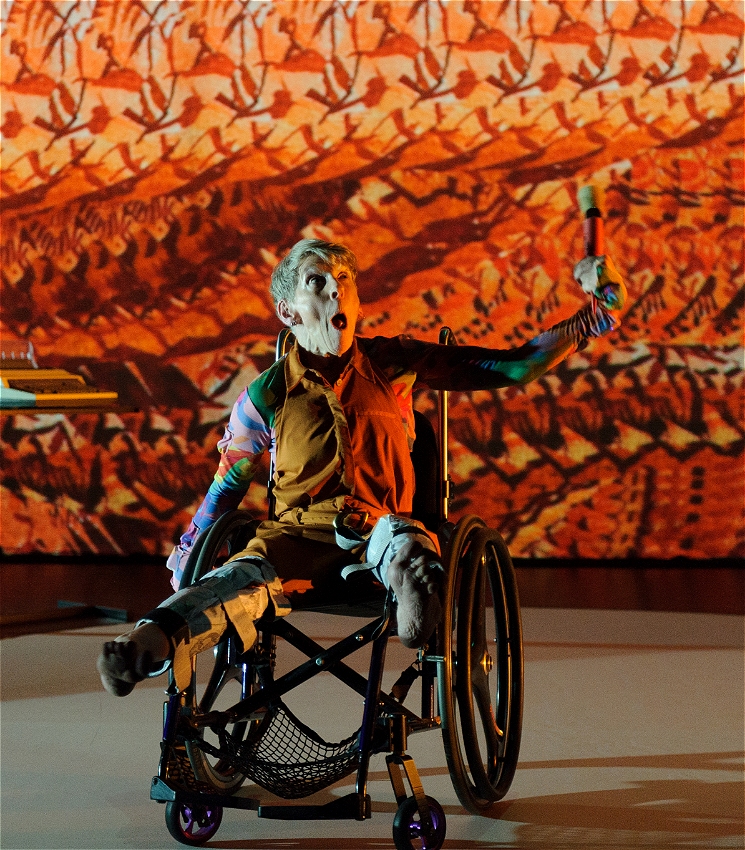An artist on stage in their wheelchair. Their mouth is open, eyes wide, and their colourful costume is partially camouflaged into the brilliantly lit background.