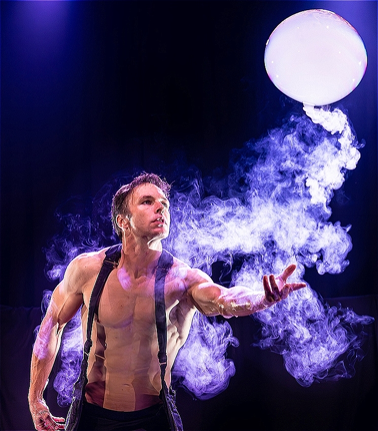 A topless man hand outstretched watches a smoke filled bubble fly and deflate as smoke surrounds him