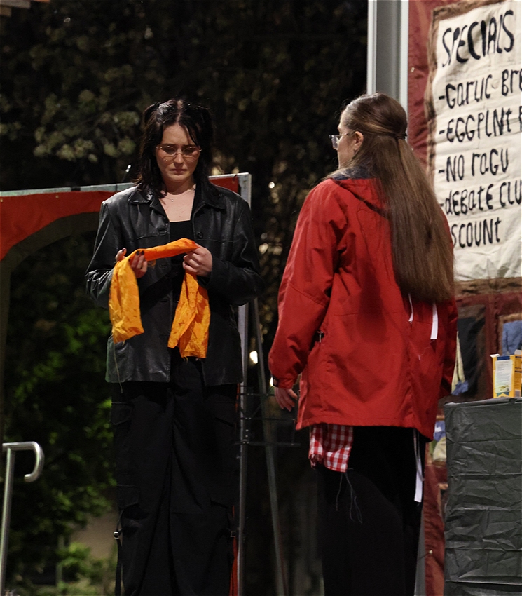 Two women stand on stage. We see one from behind, the other holds a yellow scarf, looking at it with a complicated expression..
