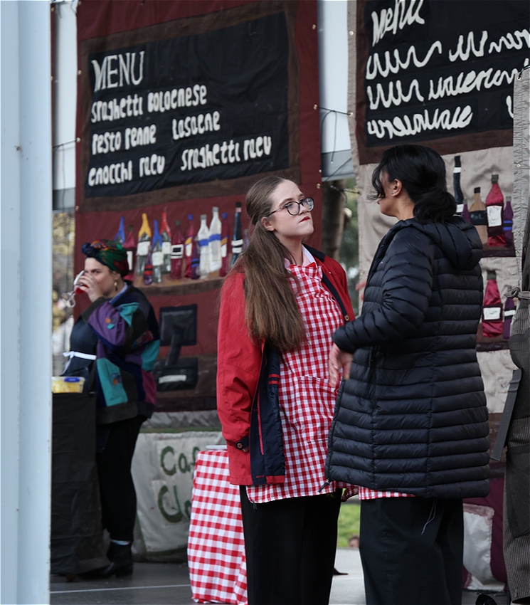 A woman in red scolds a younger girl, while a woman with a head scarf drinks, watches them from the background.
