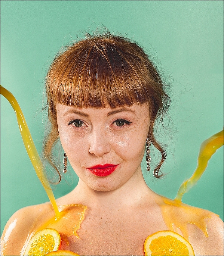 A young woman with red hair stares directly at you with a level, fiery-red-lipped, smirk. You can only see her shoulders and head, but it appears she is naked except for oranges slices on her chest. Orange juice pours from either side of her, splashing onto her shoulders.